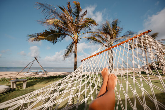 Leisure in summer . Beautiful Tanned legs of woman relax on hammock at sandy tropical beach with palm trees