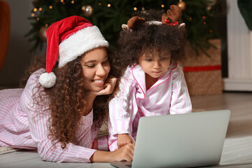 Fototapeta na wymiar Cute African-American girl and her mother with laptop at home on Christmas eve