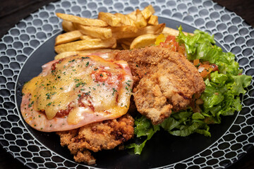 Fried chicken with french fries and sausage, cheese, salad, greens - junk food and unhealthy food. Fast food set fried chicken and french fries. Yellow sauce. Black plate on the dark wooden table.