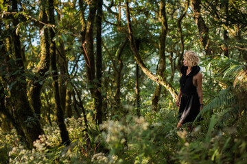 Naklejka na ściany i meble Brooding dreamy young girl blonde, black dress with a hood standing by a tree. The girl got lost in the woods. Trekking, fantasy, magical mystical forest. Girl standing by a tree. High quality photo