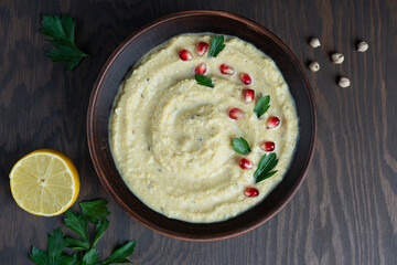 Top view of savory homemade vegetarian hummus made of chickpeas with lemon decorated with pomegranate seeds and parsley served in brown bowl on wooden background at kitchen. Horizontal orientation