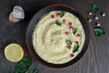 Top view of savory homemade organic hummus made of chickpeas with lemon and garlic decorated with pomegranate seeds and parsley served in brown bowl on wooden background at kitchen. Horizontal