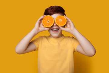 Cute ginger caucasian kid covering eye with sliced oranges posing in a t-shirt on a yellow studio wall