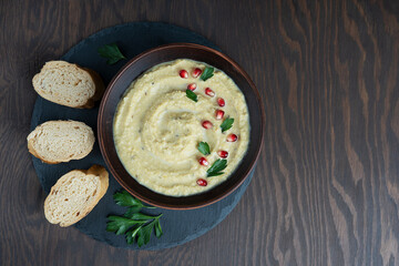 Top view of savory homemade hummus made of chickpeas  decorated with pomegranate seeds and parsley served in brown bowl on stony tray on wooden background at kitchen. Image with copy space, horizontal