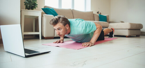 Caucasian man doing push-ups at home on the floor while using a laptop doing home based gym