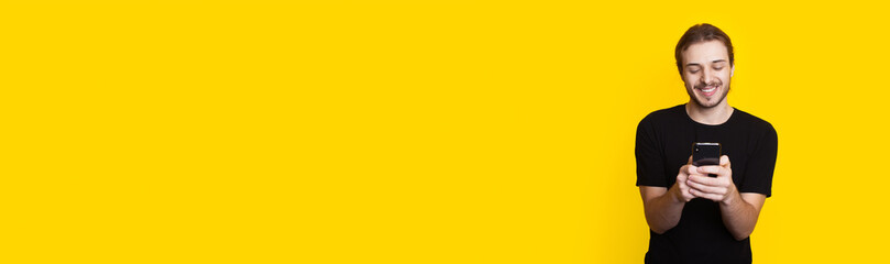 Long haired man with beard is chatting on mobile while smile in a black t-shirt on a yellow studio wall with free space