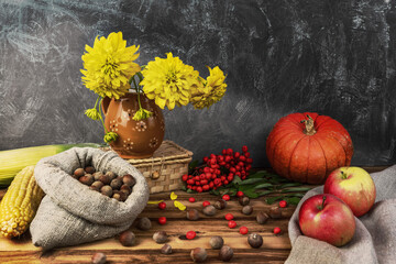 Autumn still life. Corn, apples, hazelnuts, red pumpkin, flowers in a vase, wicker box, rowan berries on a wooden background