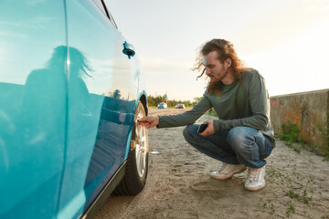 Young caucasian man checking air pressure of car tire on local road side while traveling, Male driver having troubles with his auto, checking wheel after car breakdown