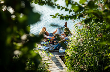 Group of friends having fun on picnic near a lake, sitting on pier eating and drinking wine.
