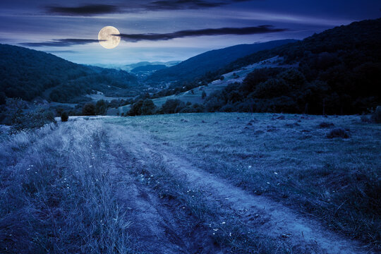 country road through rural field at night. suburban summer landscape in mountains in full moon light. village in the distant valley. cloudy day
