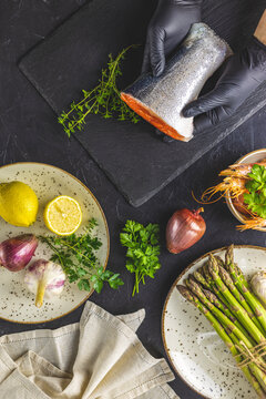 Hands in black gloves support trout fish on black stone cutting board surrounded herbs, onion, garlic, asparagus, shrimp, prawn in ceramic plate Black concrete table surface Healthy seafood background
