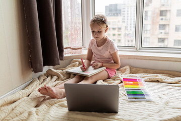Fototapeta na wymiar Little child learn english online at home. Homeschooling and distance education for kids. Caucasian Girl student drawing english letters at the notebook during coronavirus covid-19 quarantine