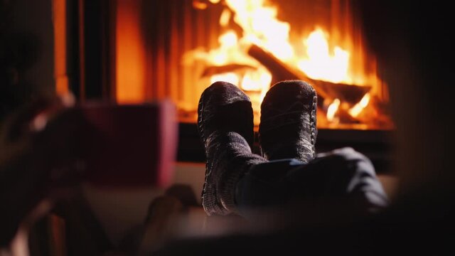 A man is resting by the fireplace, holding a cup of tea in his hand. Only socks in the frame are visible