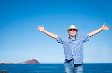 Happy old white haired man standing with outstretched arms enjoying vacation and freedom in seascape. Blue sea, red mountain and Gran Canaria island behind him