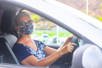 Senior woman wearing a flowered surgical mask because of the covid19 coronavirus ready to drive her gray car looking at camera - concept of active retirees