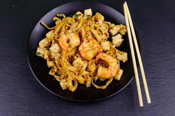Stir-fried rice noodles Pad Thai with prawns and tofu in a plate on black slate background. Top view. Thai food