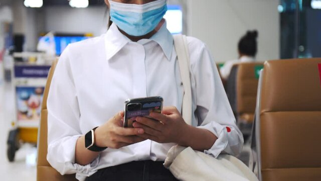Businesswoman wearing surgical mask protection and use smartphone checking social media feeds while sitting a chair in the airport terminal and keep a social distance during the COVID-19 pandemic.