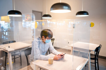 Asian man wearing protective face mask using smartphone sitting in table shield partition for preventing COVID-19 (Coronavirus) infection at cafe restaurant. Social distancing and new normal concept