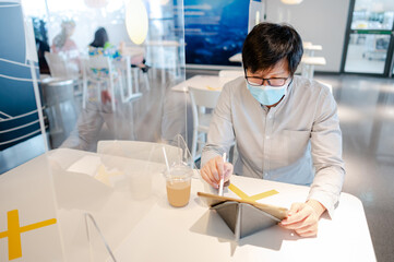 Asian man wearing face mask working with digital tablet sitting in table shield partition for preventing COVID-19 (Coronavirus) infection at cafe restaurant. Social distancing and new normal concept