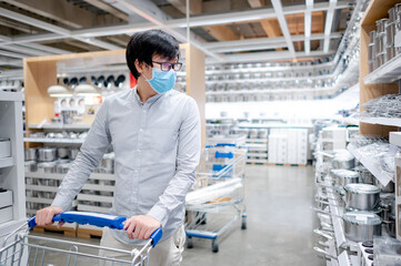 Asian man customer wearing face mask shopping in kitchenware department at grocery store or supermarket. Preventing spread of COVID-19 (Coronavirus) when buying household equipment. New normal concept