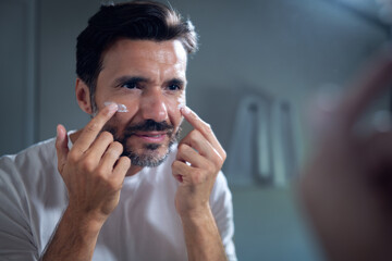 An young smiling man is applying after cleaning his face day or night cream to take care of skin behind a mirror in a bathroom.