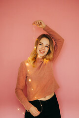 Smiling woman in sweater having fun on a pink background with Christmas lights in her hands, looking up and smiling. Isolated. Vertical.