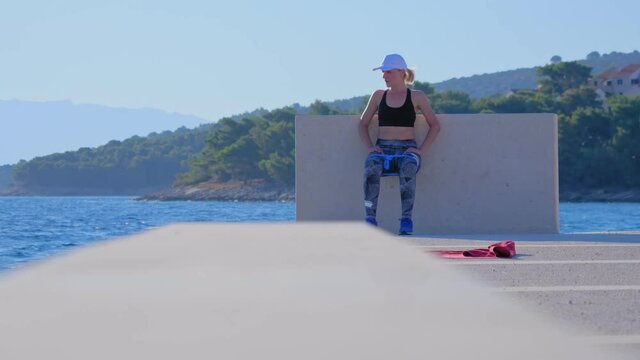 A young blonde woman in a white cap exercises outside by the sea. Wall sit exercise. A beautiful summer morning.
