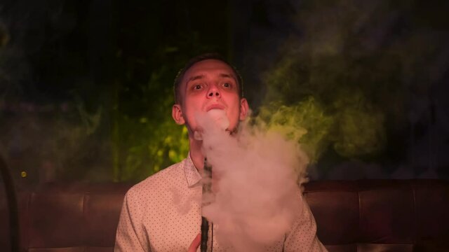 Young guy smokes hookah alone at the night bar and looking sad, concept of loneliness. Media. Blond male with bad habit, an alternative to tobacco.