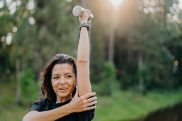 Outdoor shot of sporty European woman has dark hair raises arm with dumbbell, trains muscles and looks somewhere into distance, poses over green nature background. Female with sport equipment