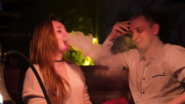 Happy couple interacting in night club while smoking shisha. Media. Young man and woman on a date talking to each other, having fun, girl exhaling clouds of smoke, concept of love.