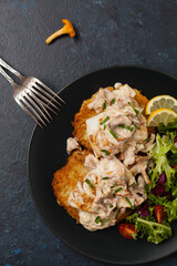 Fried cod, served on potato pancakes with mushrooms sauce and salads. Portion on a black plate. Dark background.