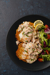 Fried cod, served on potato pancakes with mushrooms sauce and salads. Portion on a black plate. Dark background.