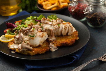 Fried cod, served on potato pancakes with mushrooms sauce and salads. Portion on a black plate. Dark background.