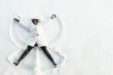 black girl in fur coat and high top boots lies on white glade and draws snow angel picture with...