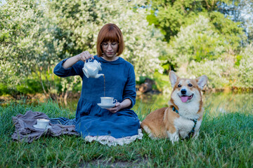 Young woman in retro dress with funny corgi dog on the picnic, female with cute dog drink english tea in the park