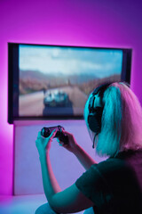 Back view shot of professional gamer playing online video game on her personal computer. Room lit in neon light and retro style. Using headphones to talk with the team. Entertainment, fun concept. © master1305