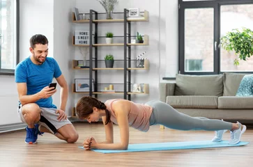 Tuinposter fitness, sport, training and lifestyle concept - happy smiling personal trainer with smartphone and woman doing plank at home © Syda Productions