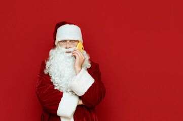 Portrait of a funny santa stands on a red background and talks on the phone with a serious face. Santa is calling on smartphone. Isolated. Christmas concept. Copyspace