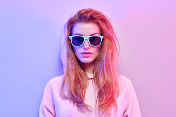 Naklejka premium Fashionable hipster woman in Trendy outfit, stylish hair, makeup on pink purple neon light. Redhead model in hoodie, fashion sunglasses. Beautiful girl in autumn fall neon style