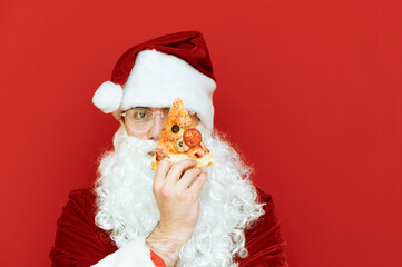 Closeup portrait of Santa Claus holding a slice of pizza in front of his eye and looking into the camera with a serious face isolated on red background. Copy space