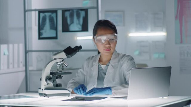 Slow motion footage of Asian woman wearing white coat, protective gloves and eyewear testing sample using microscope and laptop