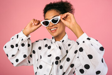 Studio photo of african black woman in stylish dress and white sunglasses. Pretty smiling female posing over pink background.
