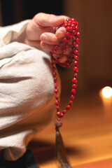Close-up of a male monk's hand holding a red wooden rosary in a dark study room. Religion faith