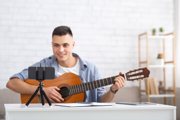 Leisure time, hobby and modern technology. Cheerful millennial man plays on guitar and looks at web camera of smartphone and studies with online teacher