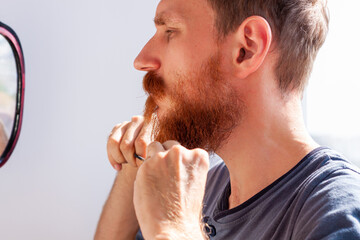 Naklejka premium Adult man cutting his own beard and mustache with scissors and comb. Caucasian red bearded male trimming hair on face at home. Do it yourself self-made haircut.Selfcare DIY during quarantine isolation