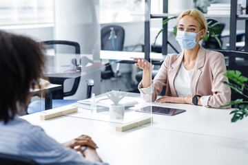 African american lady speaks to business woman in protective mask through glass partition in office...