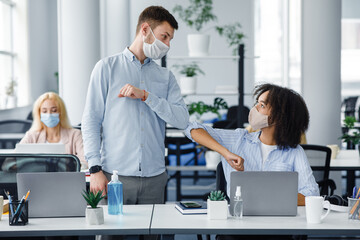 New normal and modern trend to say hello. Guy and african american lady in protective masks greet each other their elbows at workplace in interior of coworking office