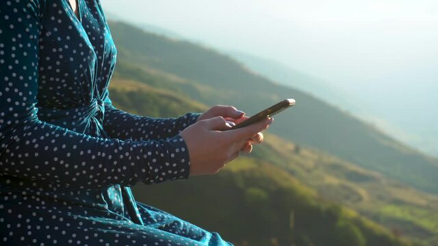 A young woman in a dress sits with a phone in her hands against a background of mountains closeup. The girl travels in the Caucasus mountains.