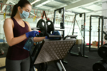 Woman cleaning exercise equipment with disinfectant spray and cloth in gym