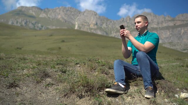 A young man sits with a phone in his hands in the mountains. The man is in correspondence of the smartphone in the Caucasian mountains.